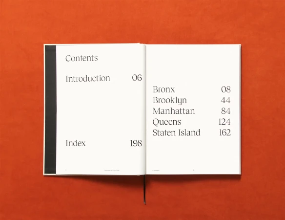 Open book on an orange surface displays 'Contents,' 'Introduction,' 'Index,' and borough names like 'Bronx' and 'Manhattan'.
