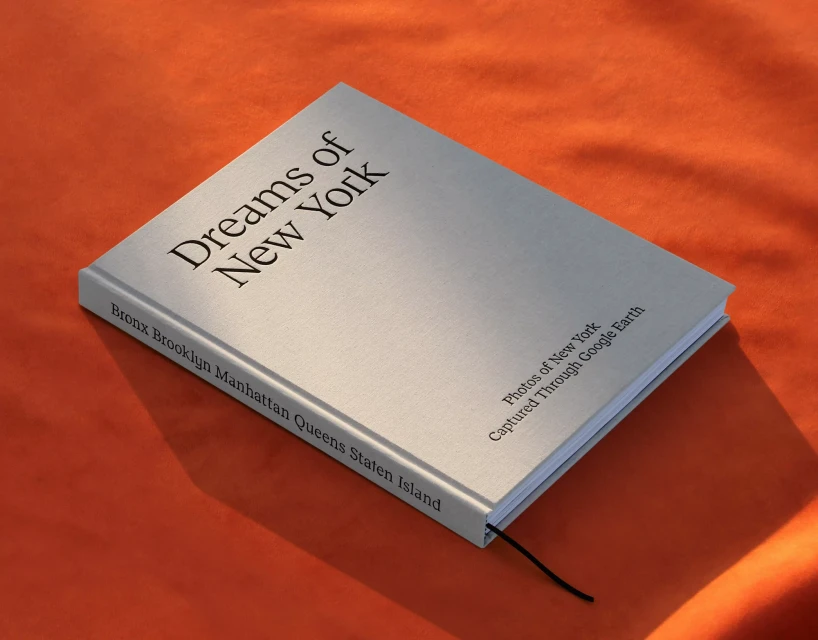 Elevated view of a book titled 'Dreams of New York' with visible text listing boroughs, captured with natural lighting on orange fabric.