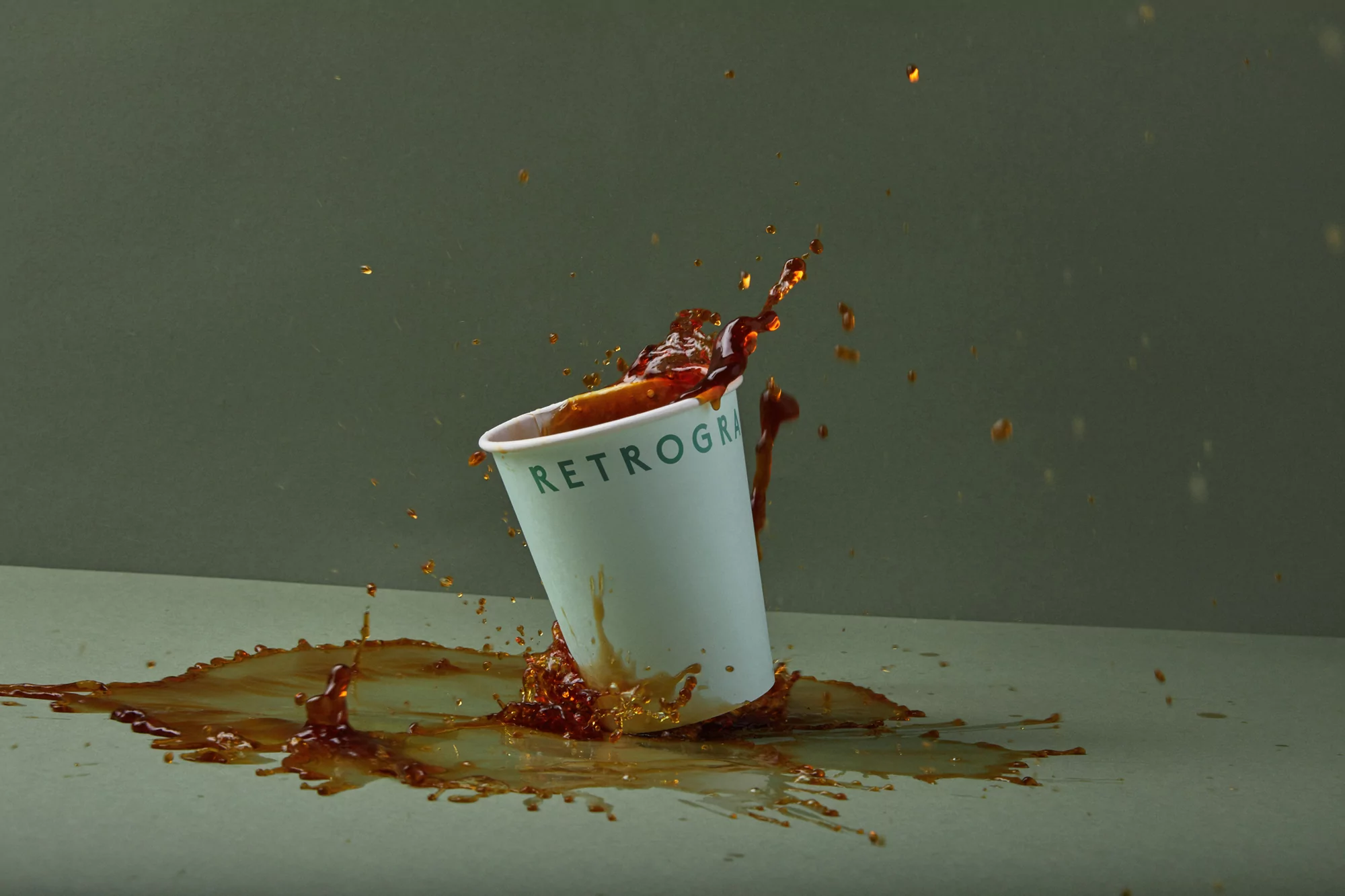 Paper cup spills a dark brown liquid onto a flat surface, creating a splash. The background is a solid greenish-gray color.