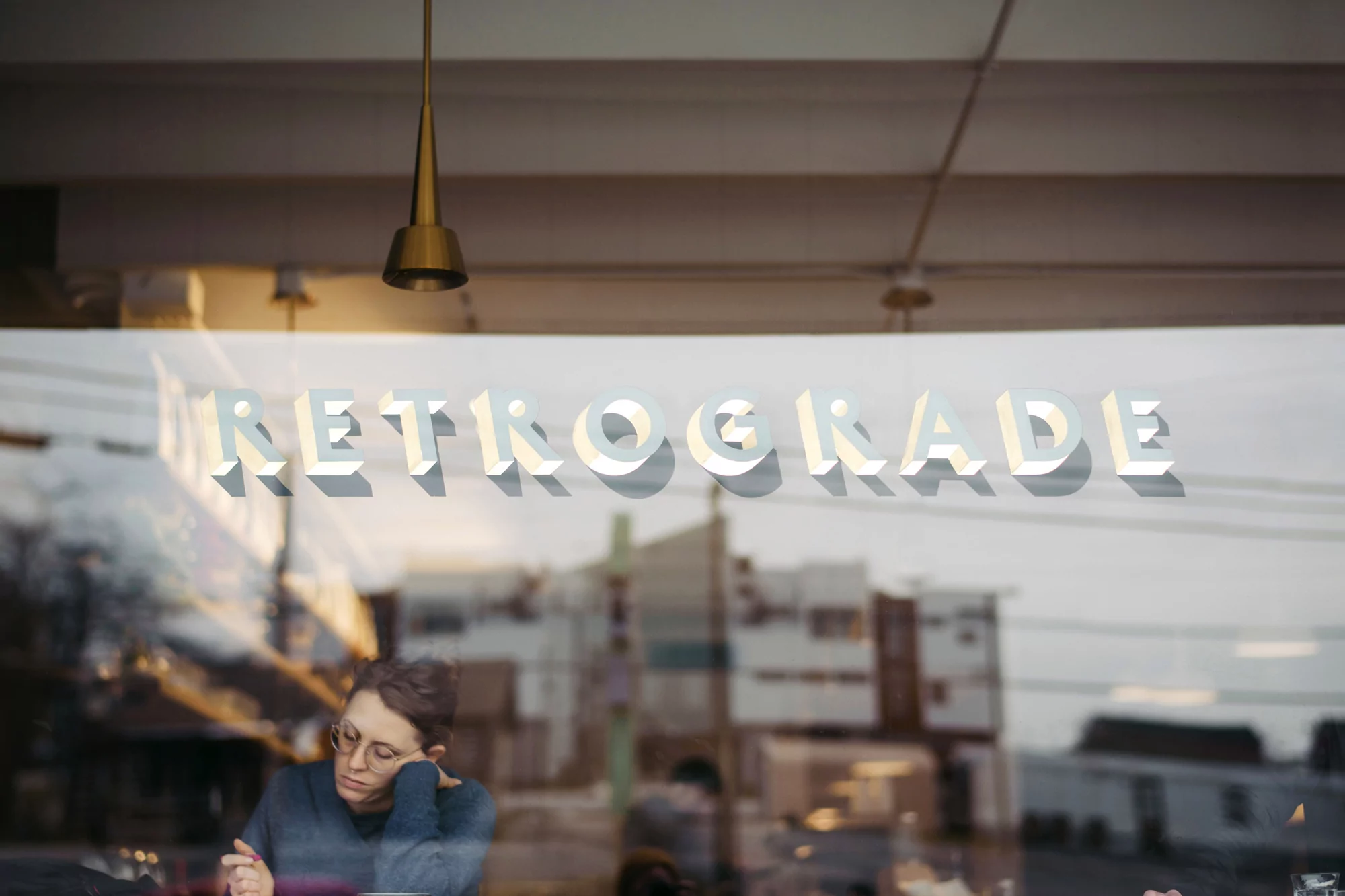 Through a window, an adult wearing glasses rests their head on their hand in a cafe, reflected with 'Retrograde' signage.