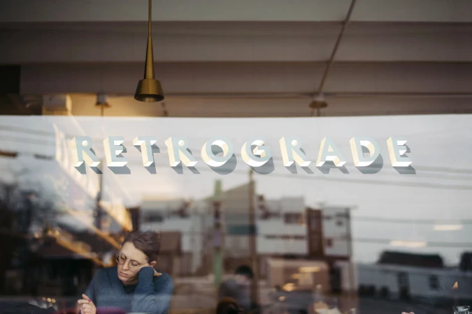 Through a window, an adult wearing glasses rests their head on their hand in a cafe, reflected with 'Retrograde' signage.