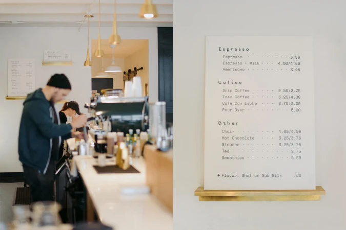 Inside a brightly lit coffee shop, an adult male barista works behind the counter as a menu board displays prices for drinks.