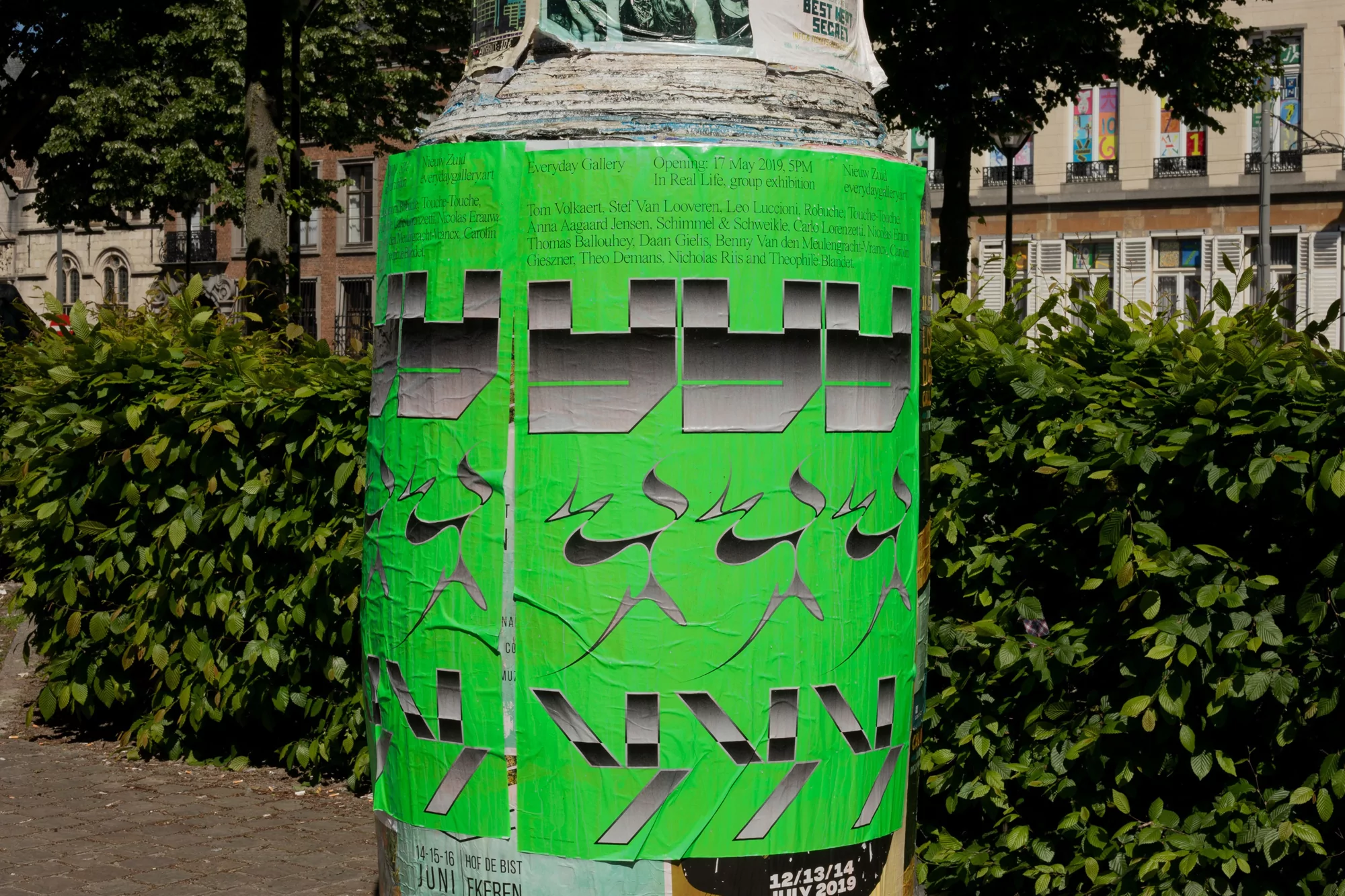 Outdoor shot of a weathered column covered in vibrant green posters with gray and black graphics, next to green bushes and a building.