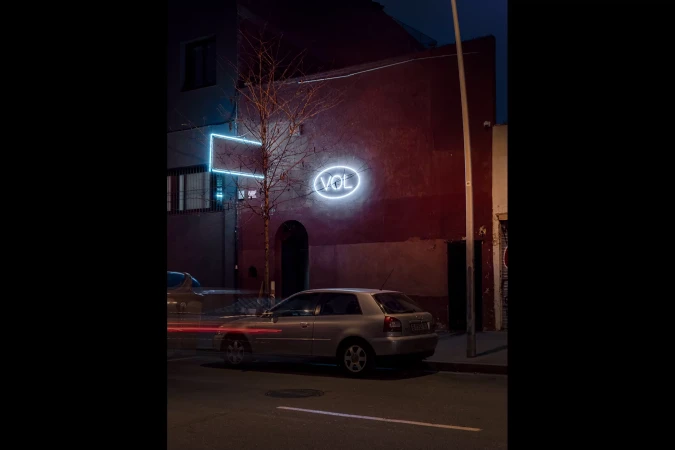 City street at night with a parked car, building with neon signs, and light trails from passing vehicles adding dynamic movement.