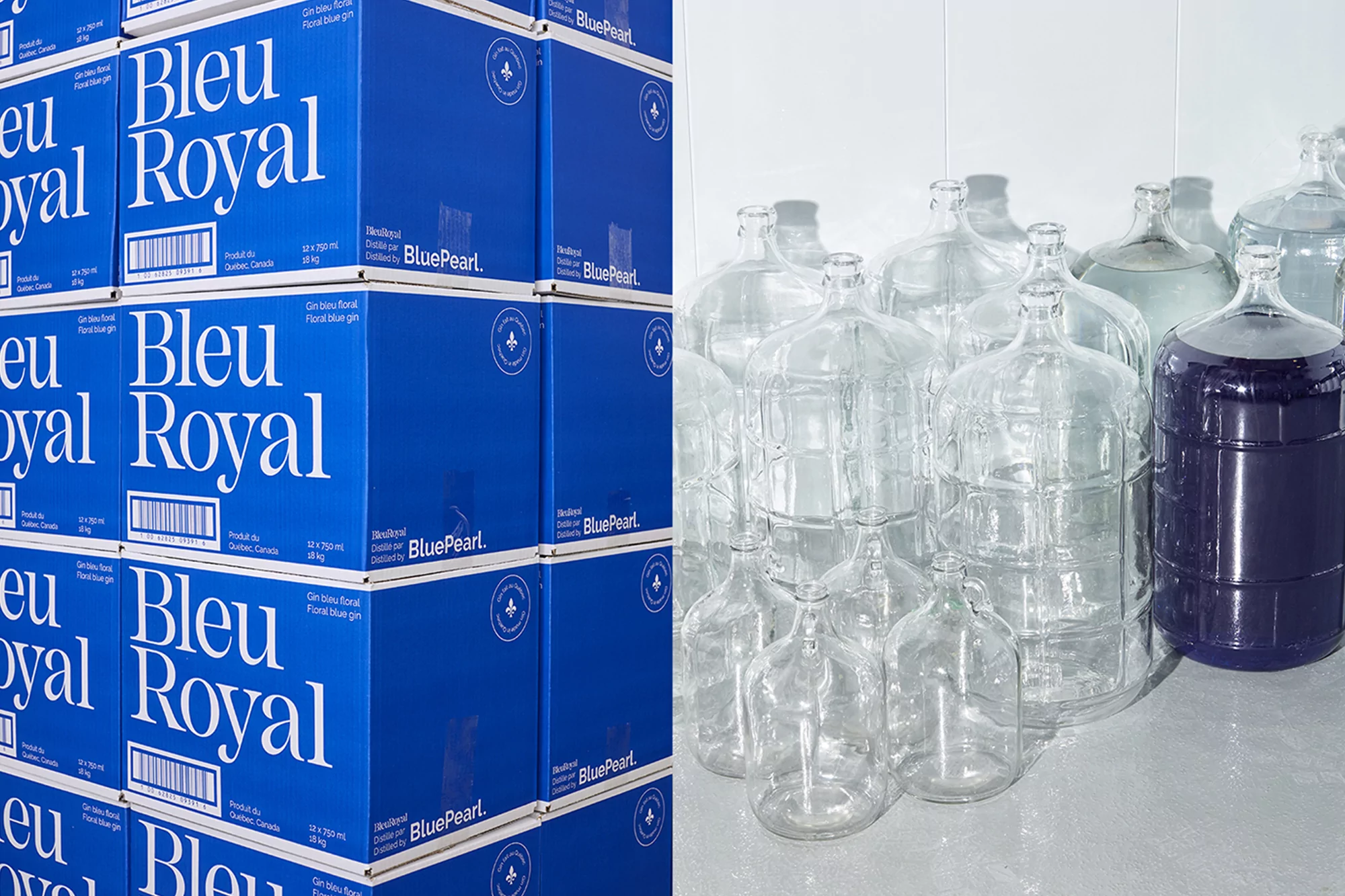 Stacked blue boxes with gin brand name beside clear and blue liquid filled carboys on a bright background in a studio setting.