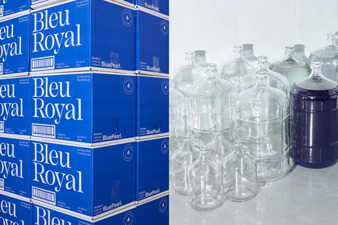 Stacked blue boxes with gin brand name beside clear and blue liquid filled carboys on a bright background in a studio setting.