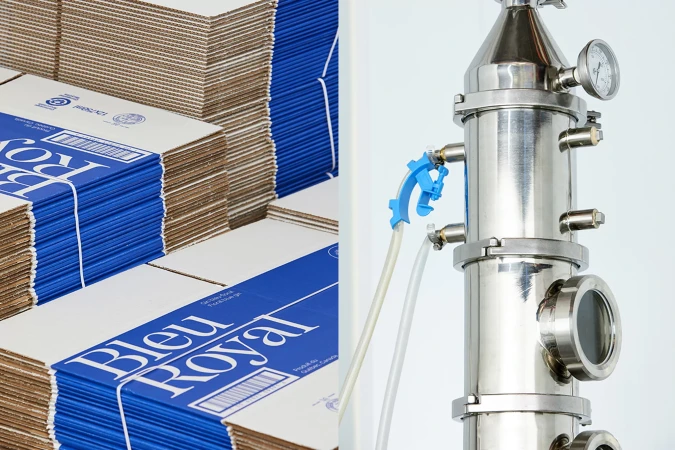 Stacks of white and blue branded cardboard boxes next to a stainless steel distiller column with tubes, gauges, and a bright background.