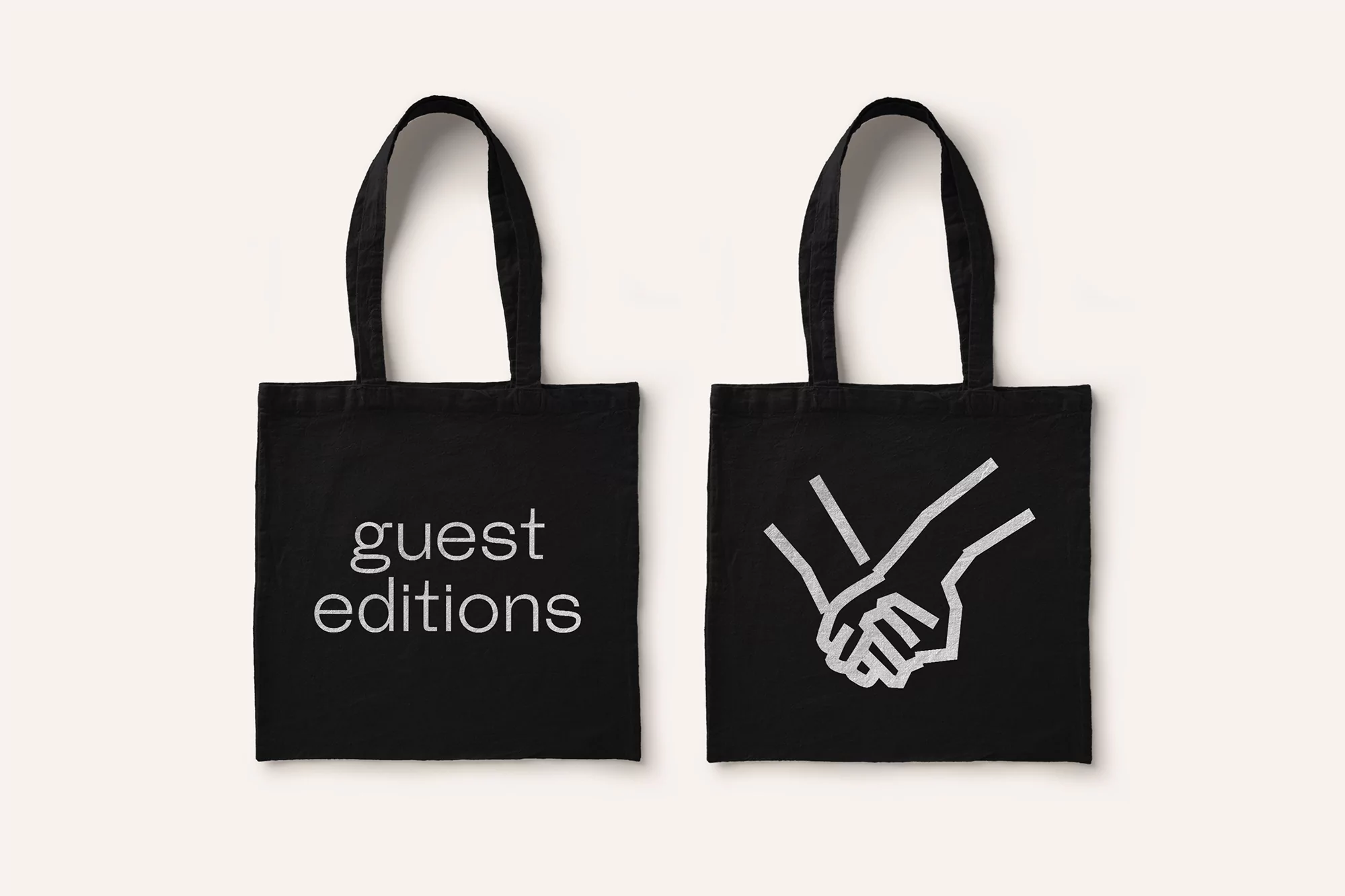 Two black tote bags sit on a cream-colored background, one featuring the words 'guest editions' and the other displaying an illustration of hands.