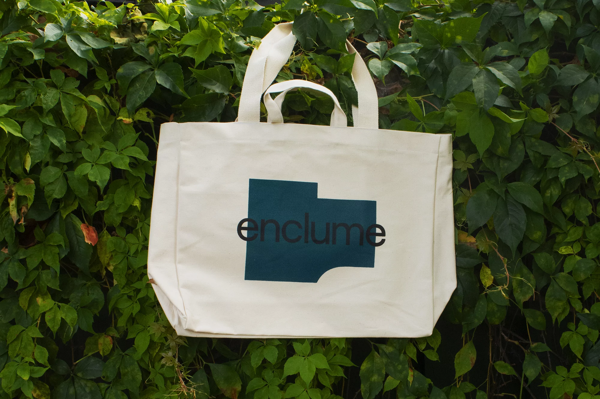Cream canvas tote bag with black and dark blue typography logo against a dense background of green leaves.
