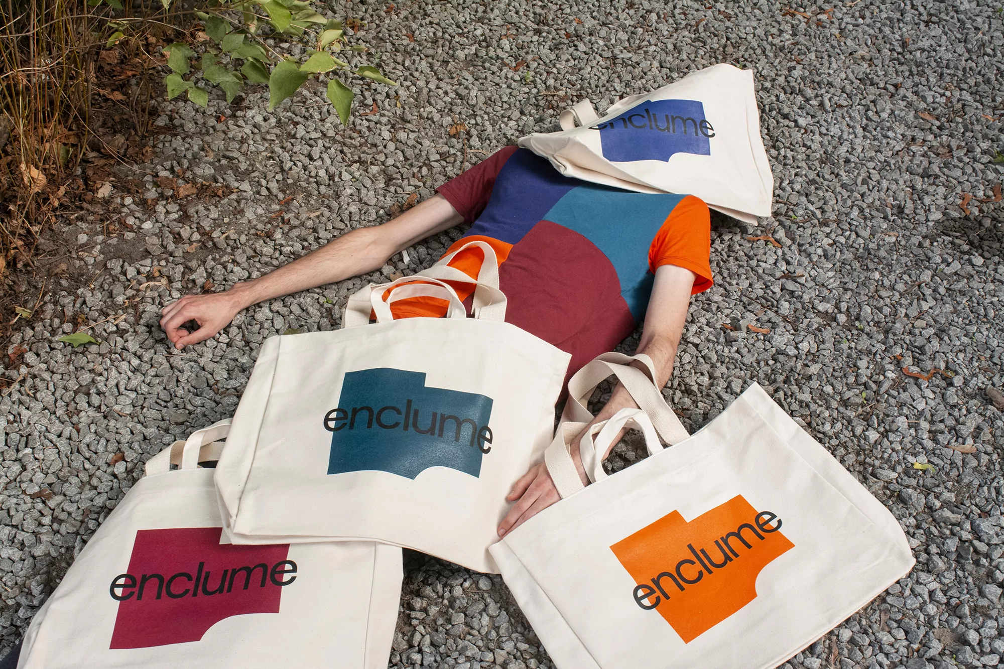 Adult lies on gravel surrounded by branded tote bags in outdoor setting. Person wears color-block t-shirt in burgundy, orange, teal, and blue.