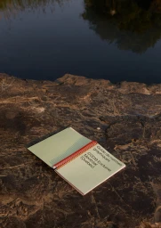 Open spiral-bound notebook resting on a dark rocky surface near a body of water displaying text on the cover