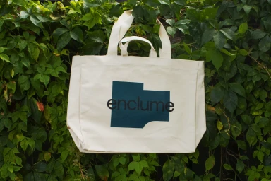 Cream canvas tote bag with black and dark blue typography logo against a dense background of green leaves.