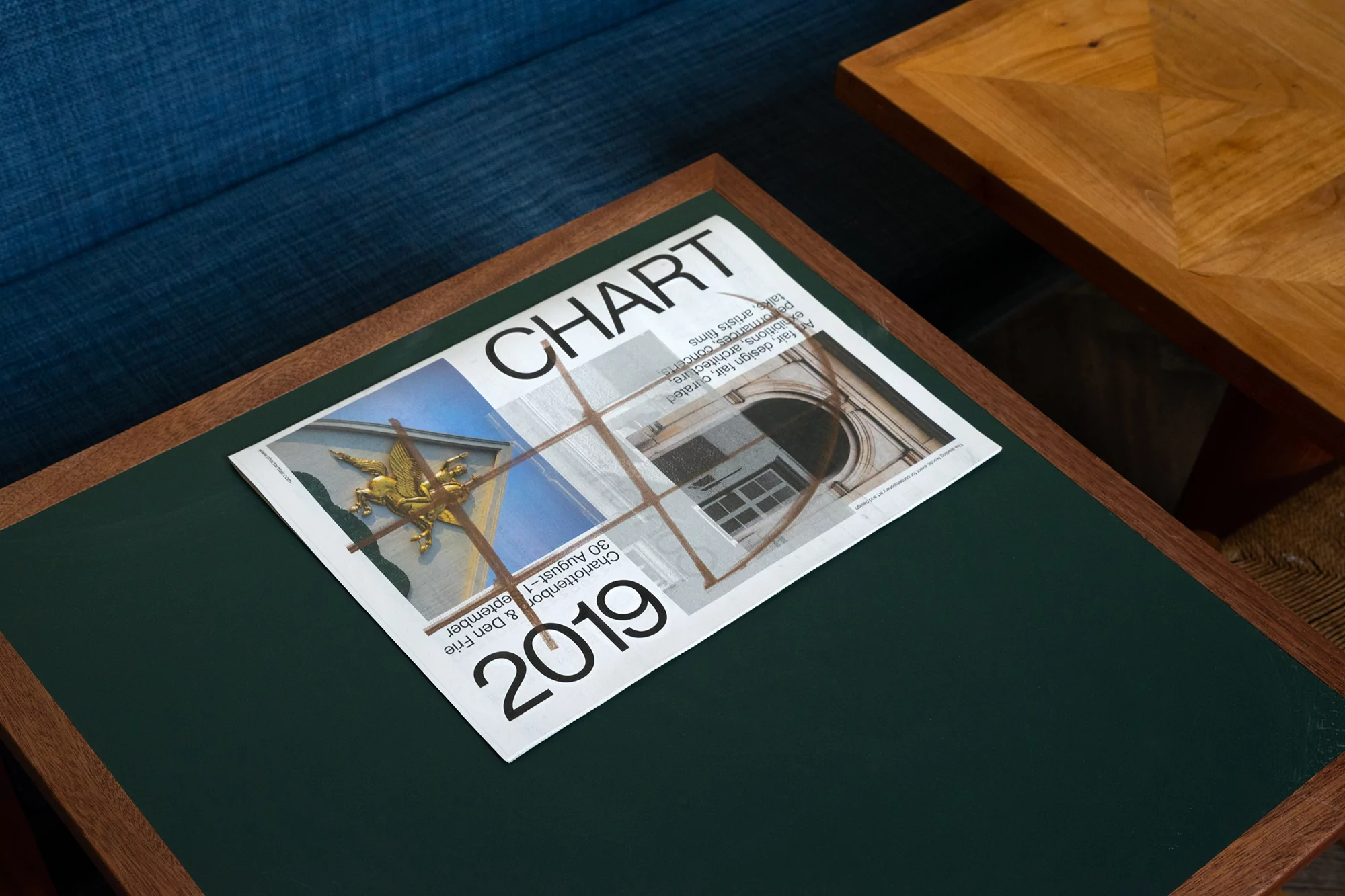 Close-up of an open magazine on a dark green table with a wood frame, next to another wooden table and a blue sofa in the background.