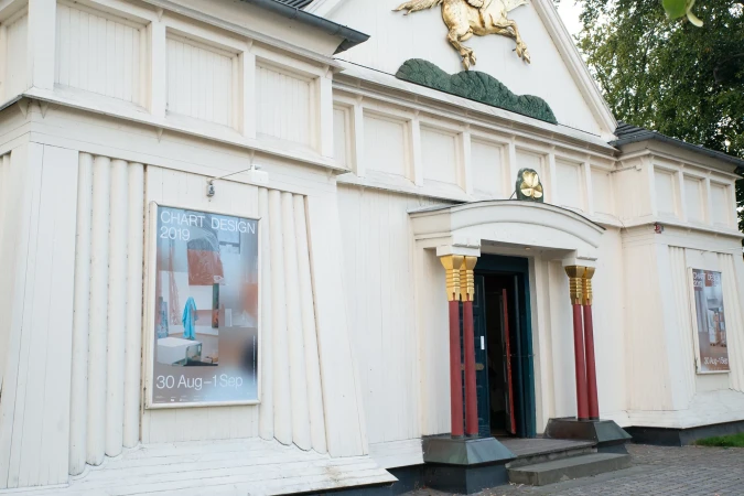 Exterior shot of a white building with a poster advertising 'Chart Design 2019' and red columns framing the open entrance.