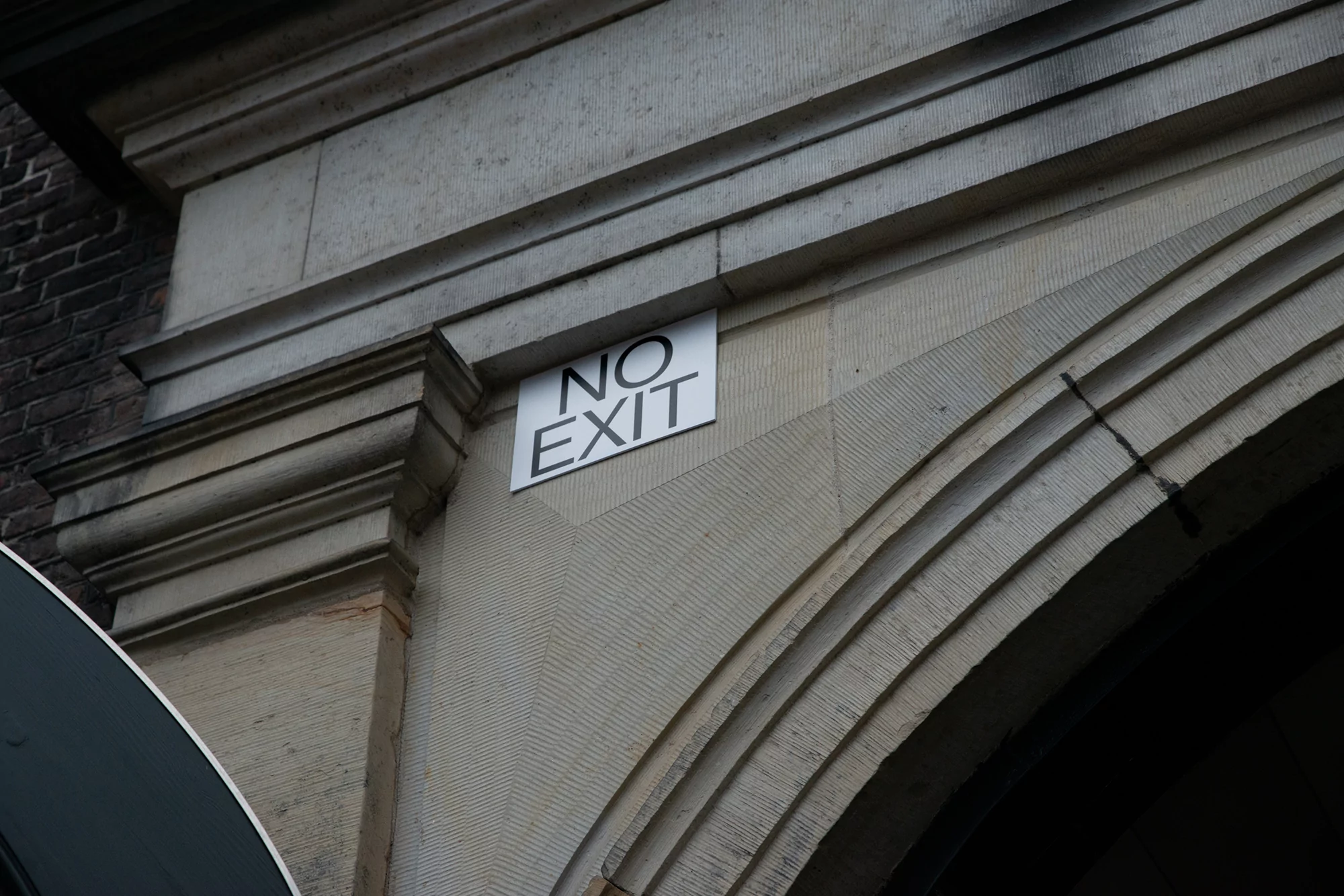 Close-up of a 'No Exit' sign mounted on a textured beige stone wall, forming part of an arched architectural structure.