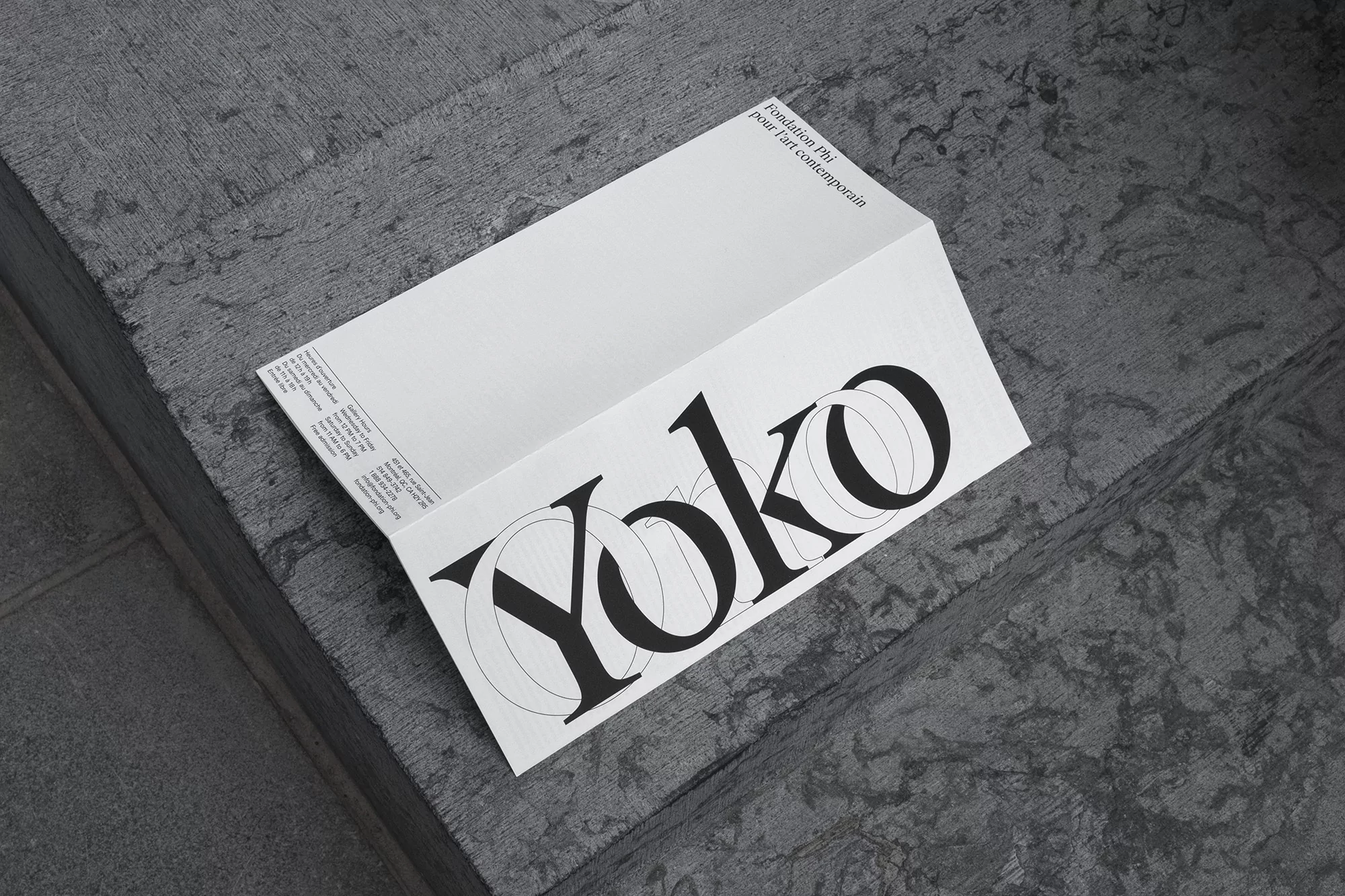 Promotional material featuring a white folded paper on textured grey concrete, showcasing bold black typography and minimalist design.