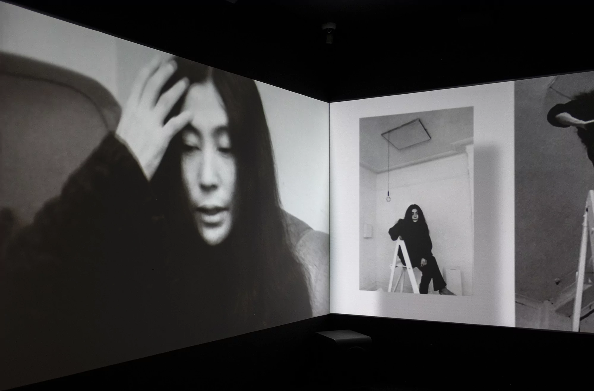 Black and white video installation features a woman's face and a woman sitting on a ladder in a white room in a dark setting.