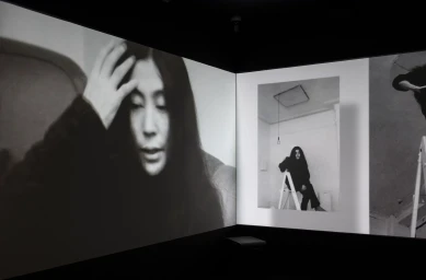 Black and white video installation projecting images of Yoko Ono across corner walls in a dark gallery space