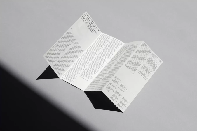 A folded printed brochure with typography resting on a plain grey surface casting a sharp dark shadow