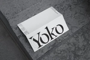 Folded white paper brochure with Yoko Ono branding featuring black typography resting on a grey stone surface.