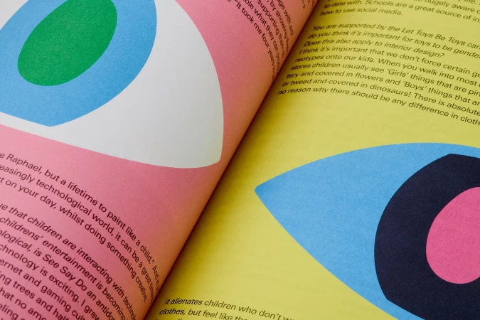 Open magazine with geometric eye illustration in pink, green, blue, and black, next to articles and columns of printed text on yellow and pink pages.