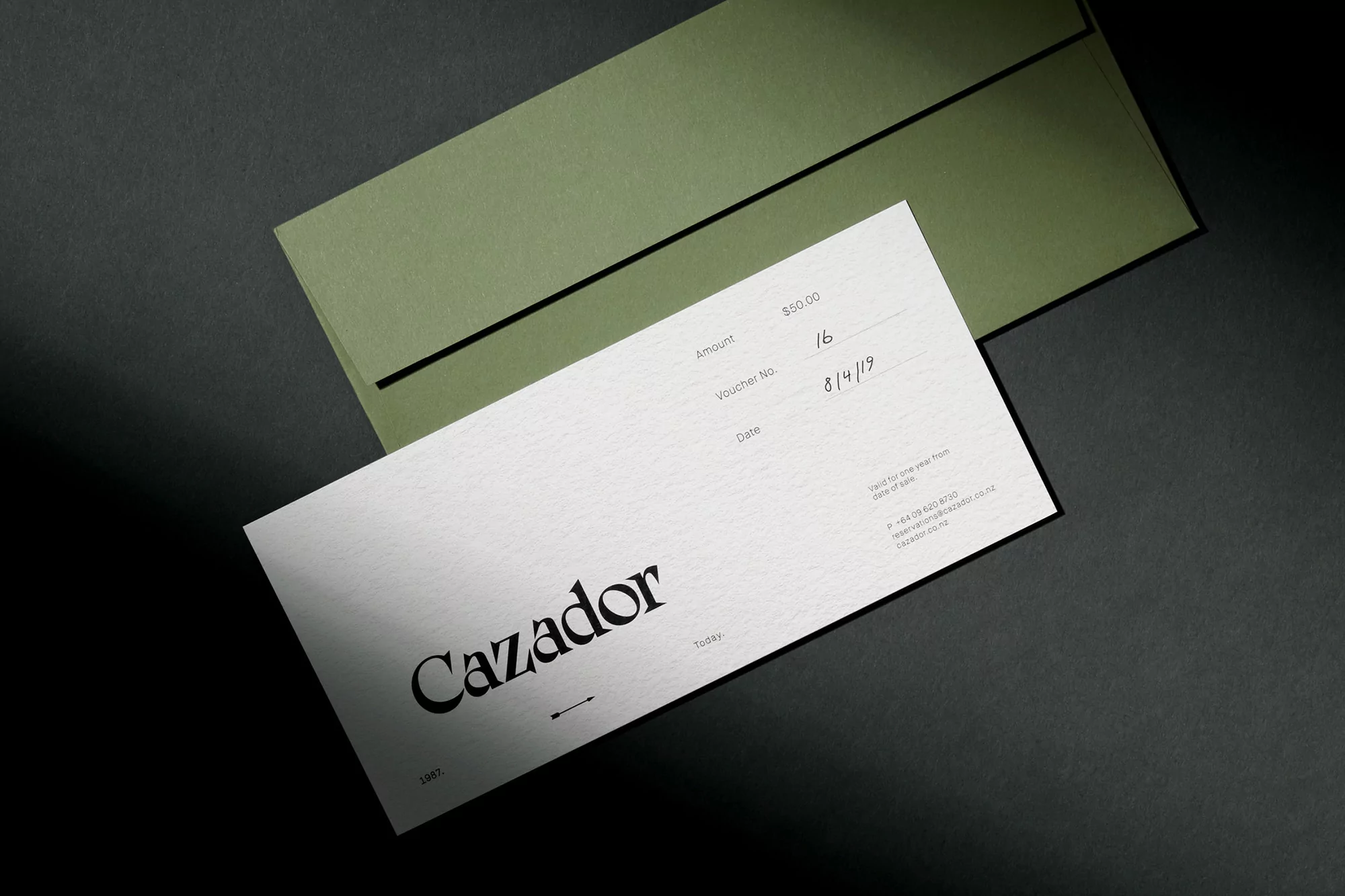 Olive green envelope and white voucher or document with black modern typeface displayed on dark gray surface, minimal graphic design.