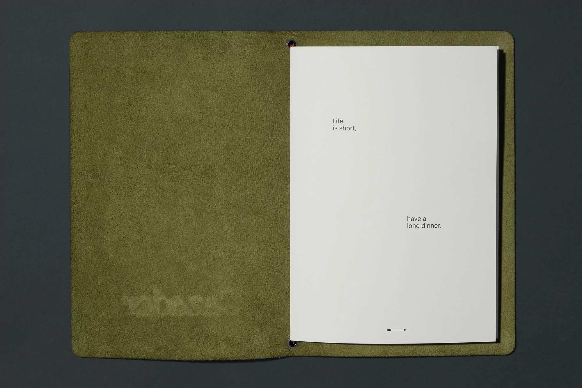 Open journal with a green leather cover on a gray surface, featuring the text 'Life is short, have a long dinner' inside.