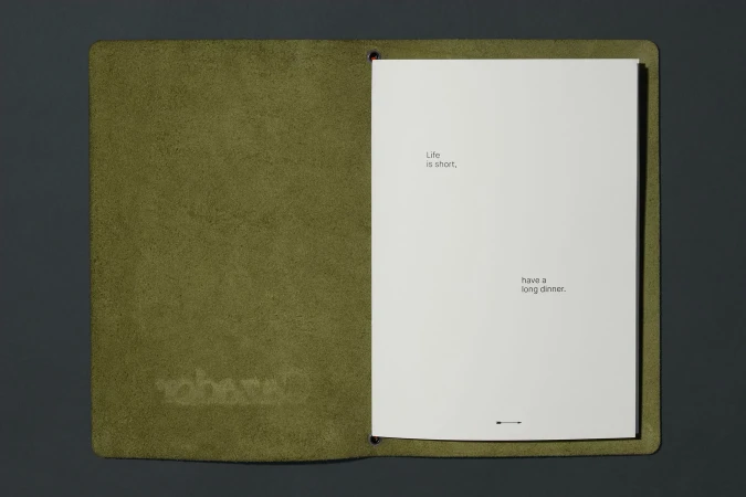 Open journal with a green leather cover on a gray surface, featuring the text 'Life is short, have a long dinner' inside.