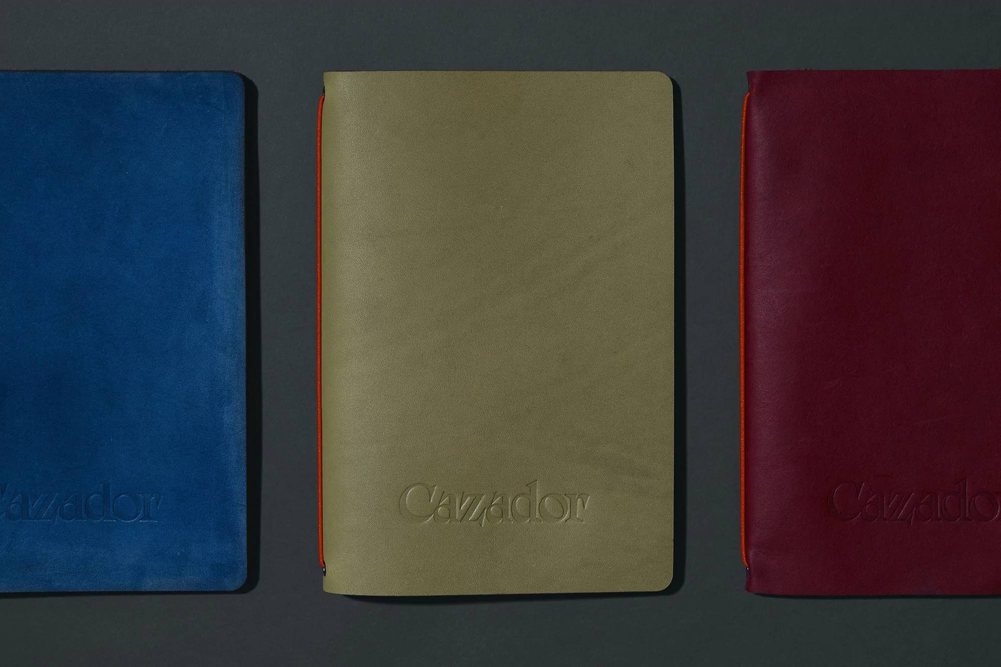 Three colorful 'Cazador' notebooks: a blue, a khaki green, and a dark red one, displayed side-by-side against a solid grey backdrop.
