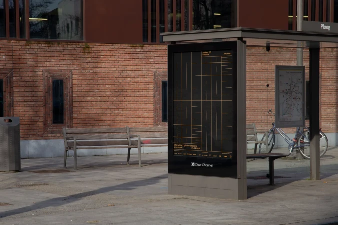 Bus stop on city street featuring a digital advertisement display, bench seating, a bicycle, and a brick building facade.