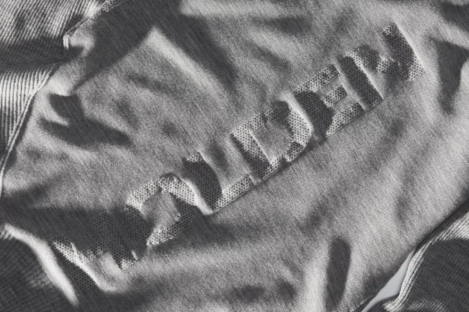 Close-up of a heather grey cotton sweater or sweatshirt with a raised, textured word design, lit by soft, indirect light.