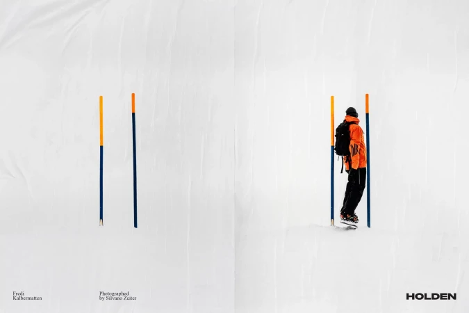 Back view of a man in orange jacket skiing between blue and orange poles on a sunny, snow-covered surface with a stark white background.