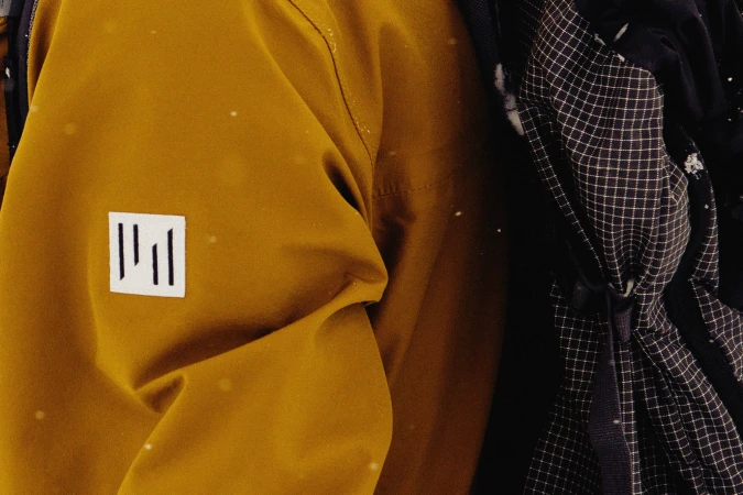 Close-up of a golden-brown jacket with a logo patch, a black backpack with white checkered pattern, and bokeh effect from snowfall.
