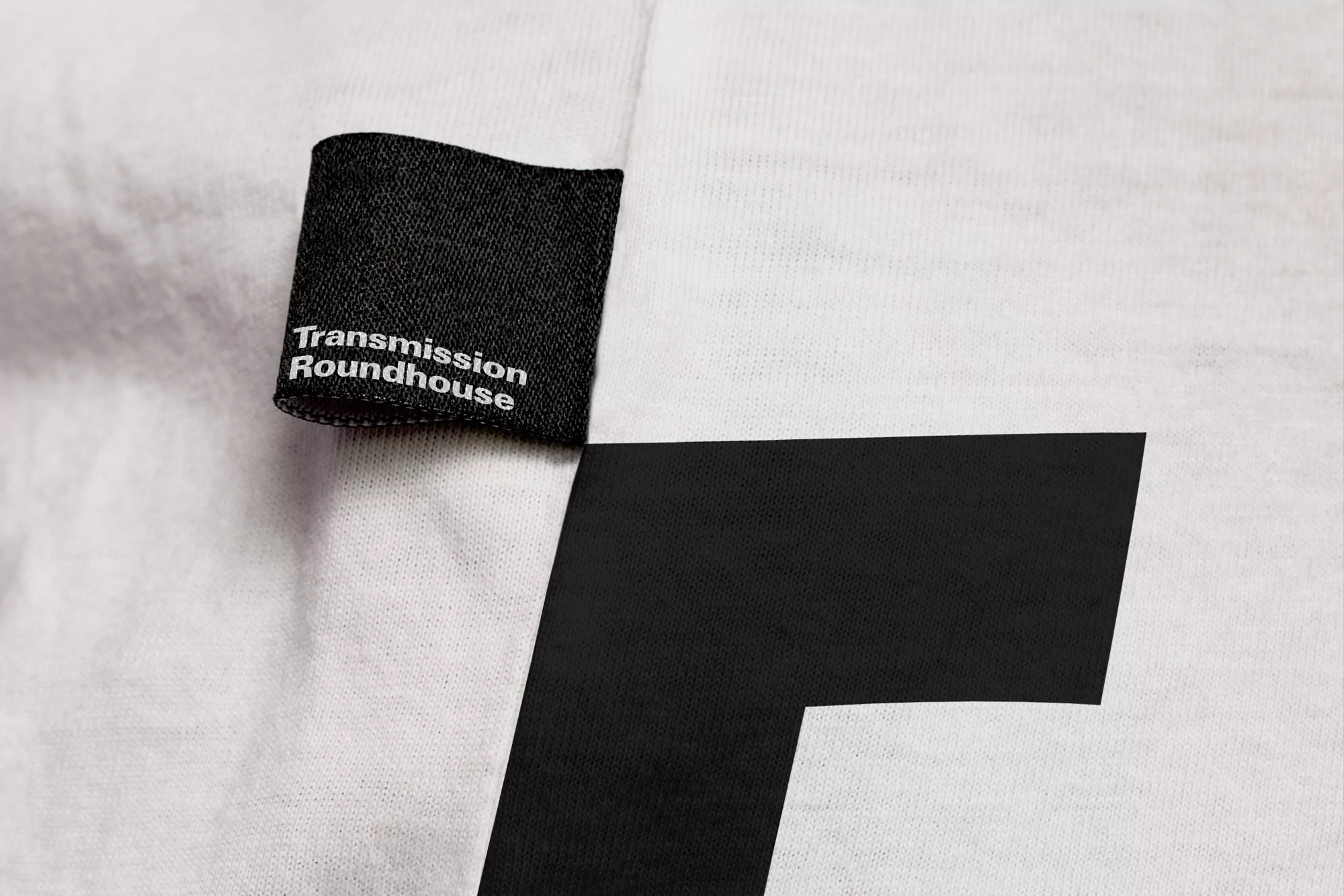Close-up of a white fabric surface with a black rectangular label stitched on the side, displaying white text 'Transmission Roundhouse'.