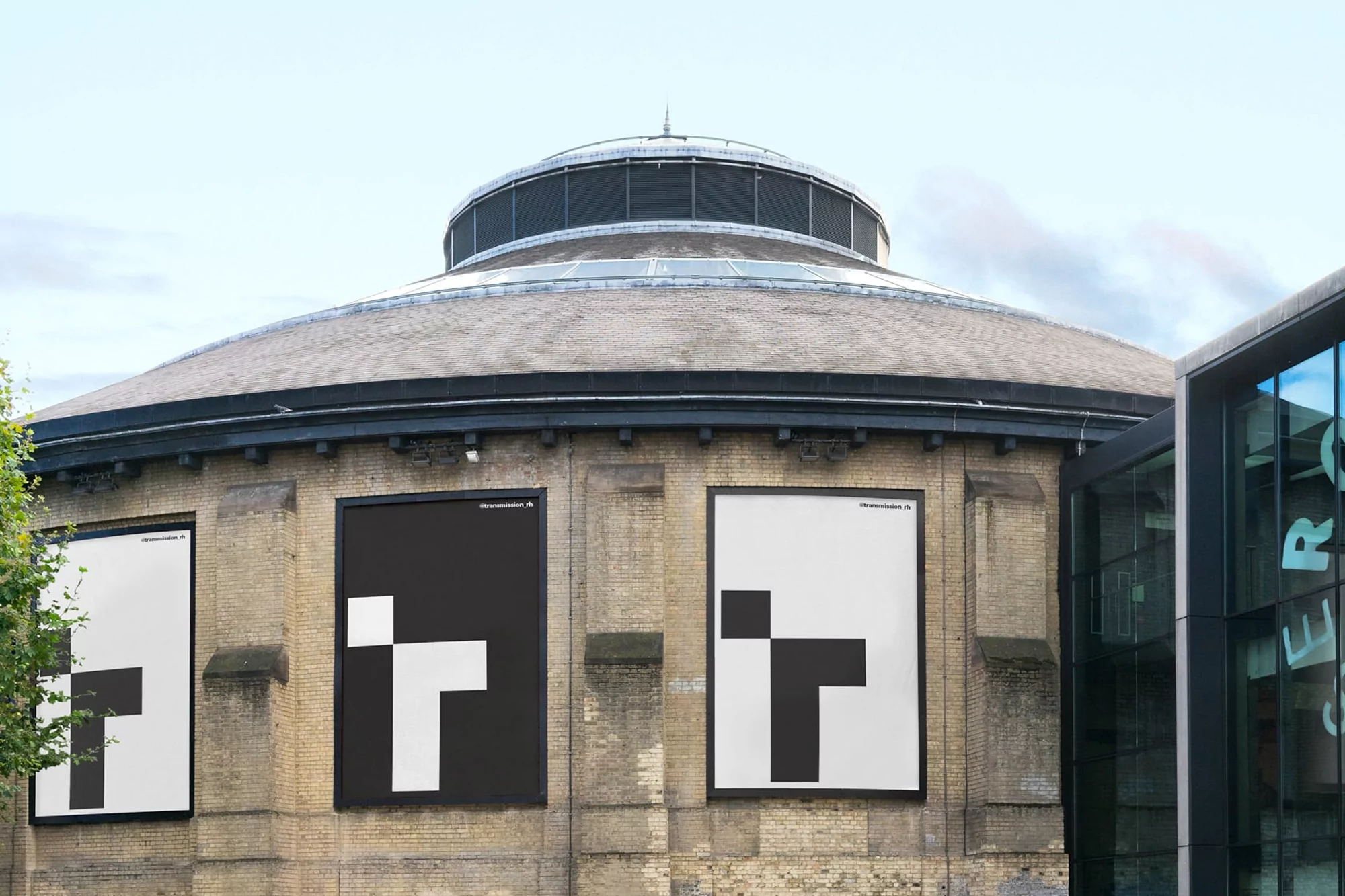 Three vertical posters with a graphic T design are mounted on the exterior brick wall of a cylindrical building under a bright sky.
