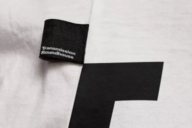 Close-up of a black fabric clothing tag with white typography reading Transmission Roundhouse attached to white fabric.
