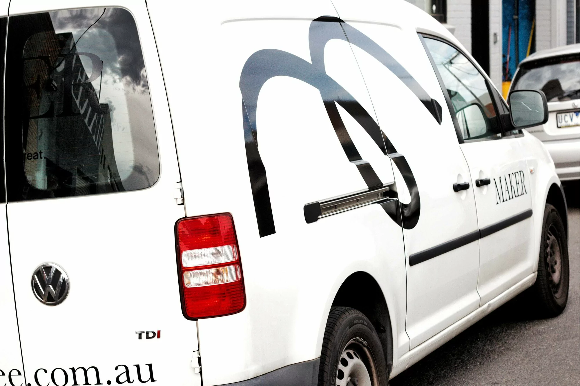 White Volkswagen van features a large black abstract logo, "MAKER" text, and a red taillight, captured in an urban outdoor setting.