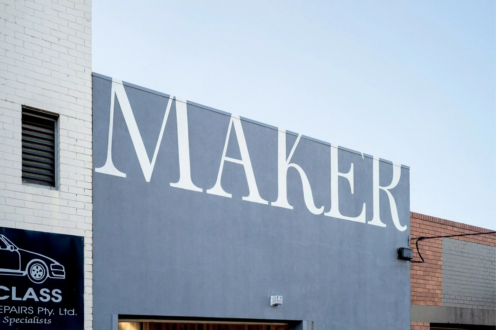 Exterior view of a building with 'MAKER' signage in white letters on a grey facade, featuring a white brick section and blue sky.