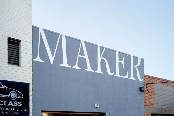 Exterior view of a building with 'MAKER' signage in white letters on a grey facade, featuring a white brick section and blue sky.