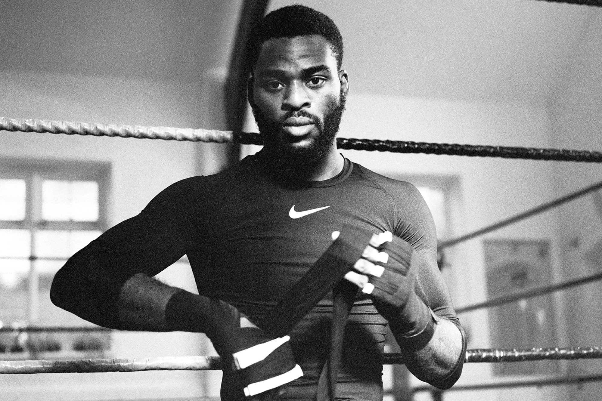 Monochrome portrait of a muscular Black male boxer in a long-sleeve shirt with hand wraps leaning against the ropes in a boxing ring.