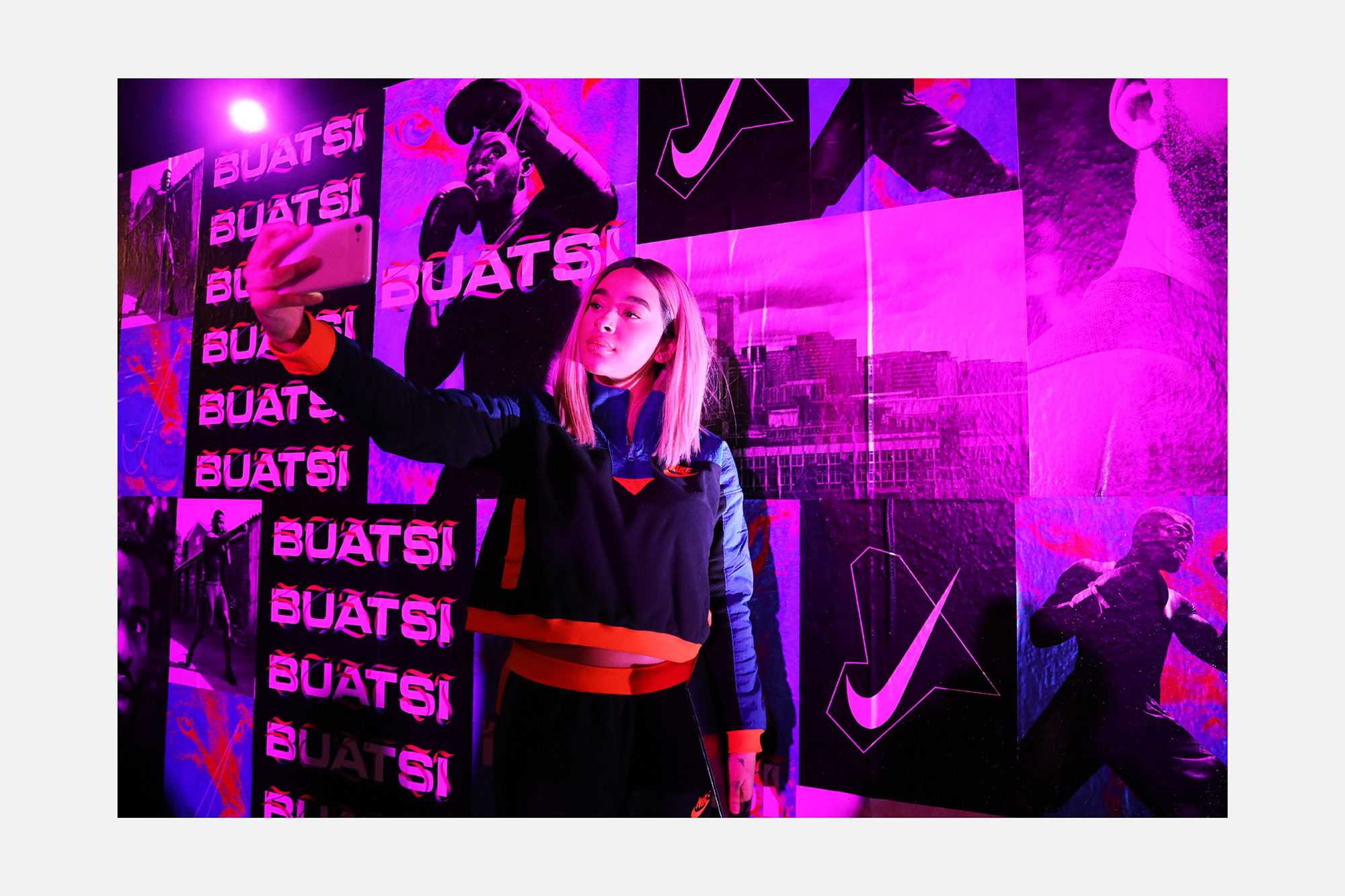 Woman in athletic wear takes a selfie indoors against a backdrop of athletic poster art in vibrant shades of pink and blue.