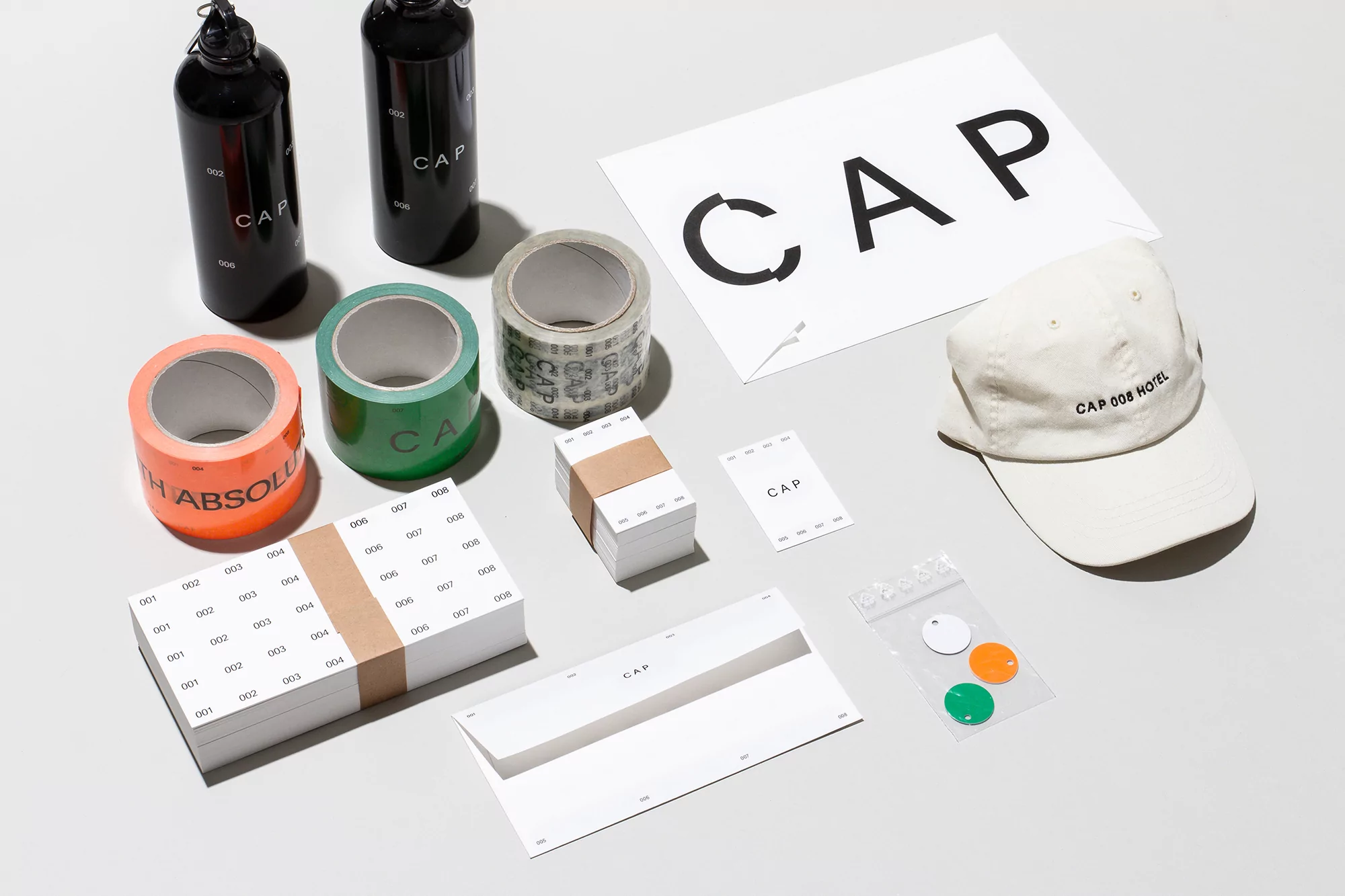 Still life of paper goods including a baseball cap, water bottles, tape rolls, envelopes, and stickers, all labelled with the word CAP.