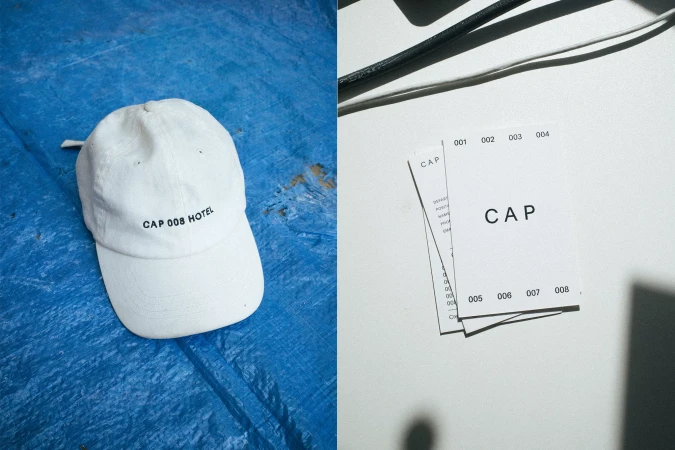 White baseball cap with 'CAP 008 HOTEL' text on a blue textured surface, alongside cards with 'CAP' printed, against a bright white backdrop.