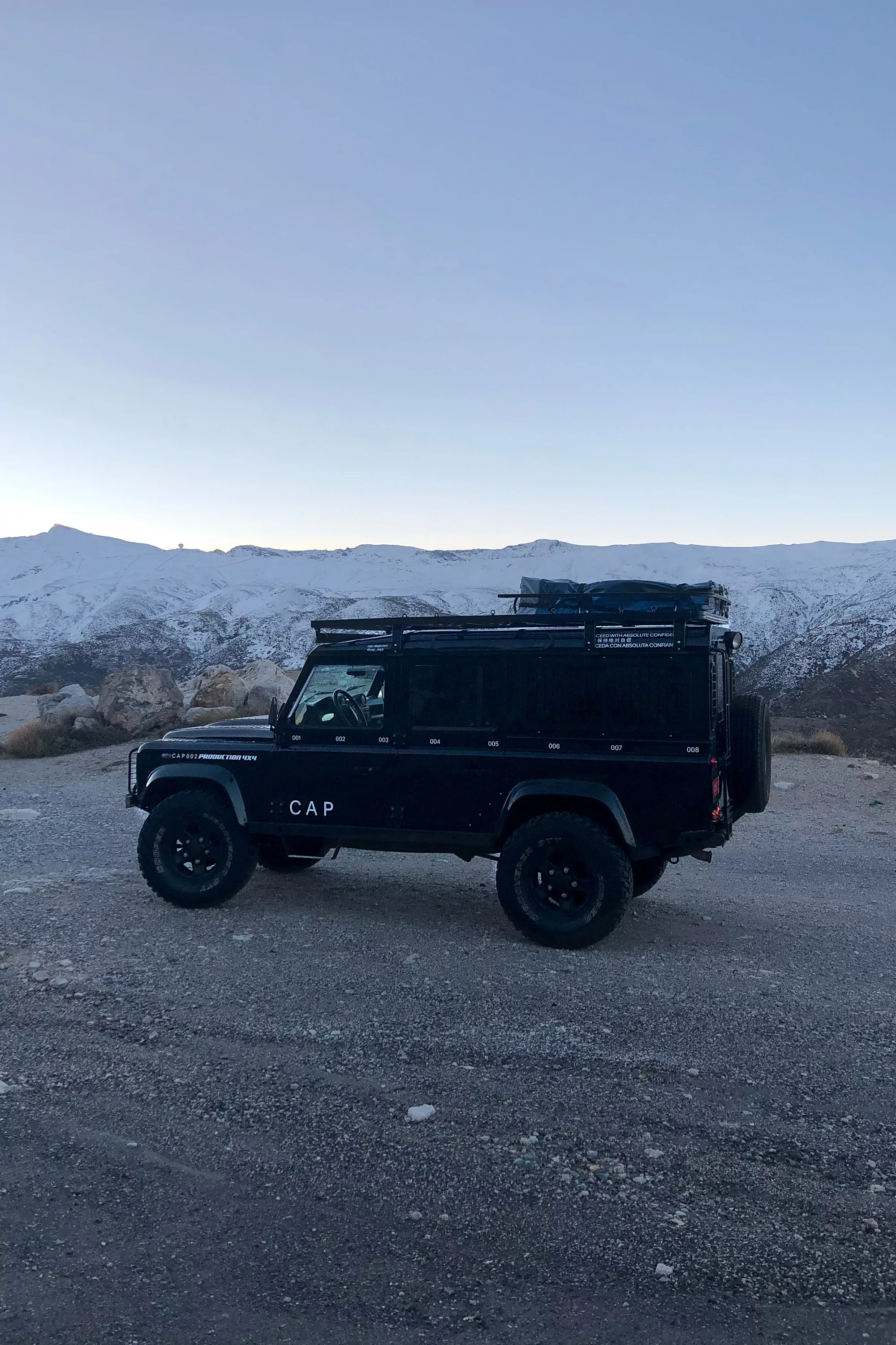 Black Land Rover Defender parked on a gravel surface, snowy mountains in background under a clear sky, captured in a side view.