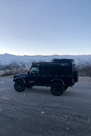 Black Land Rover Defender parked on a gravel surface, snowy mountains in background under a clear sky, captured in a side view.