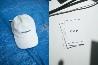 White baseball cap with black text resting on a blue textured surface alongside printed cards on a white table.