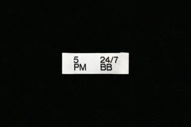 Small white label with black sans-serif typography reading 5 PM and 24/7 BB centered on a solid black background.
