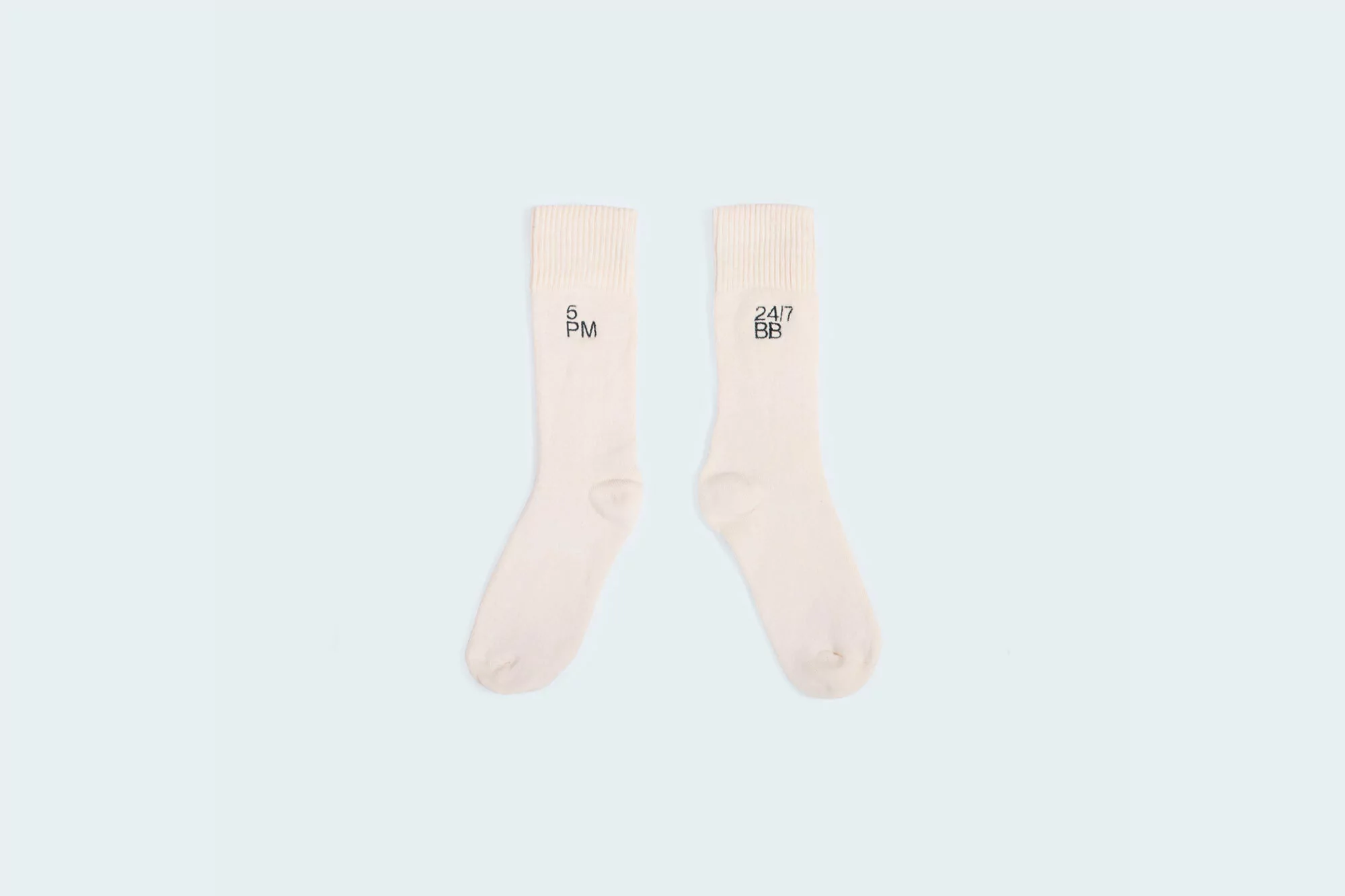 Pair of cream-colored socks with black lettering reading '5 PM' and '24/7 BB' on a light blue background in a studio shot.