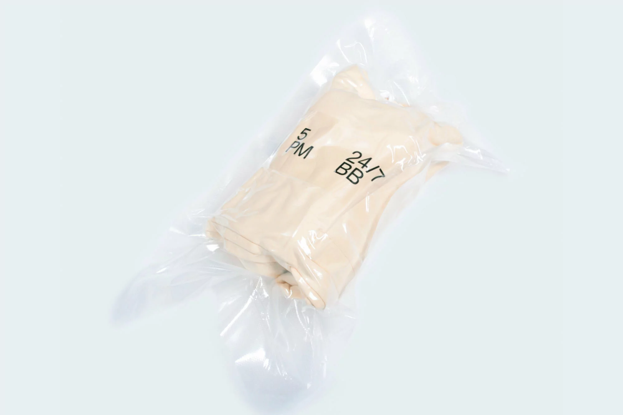 Beige clothing item in a clear plastic package, isolated on a white background, featuring '5 PM' and '24/7 BB' printed on it.