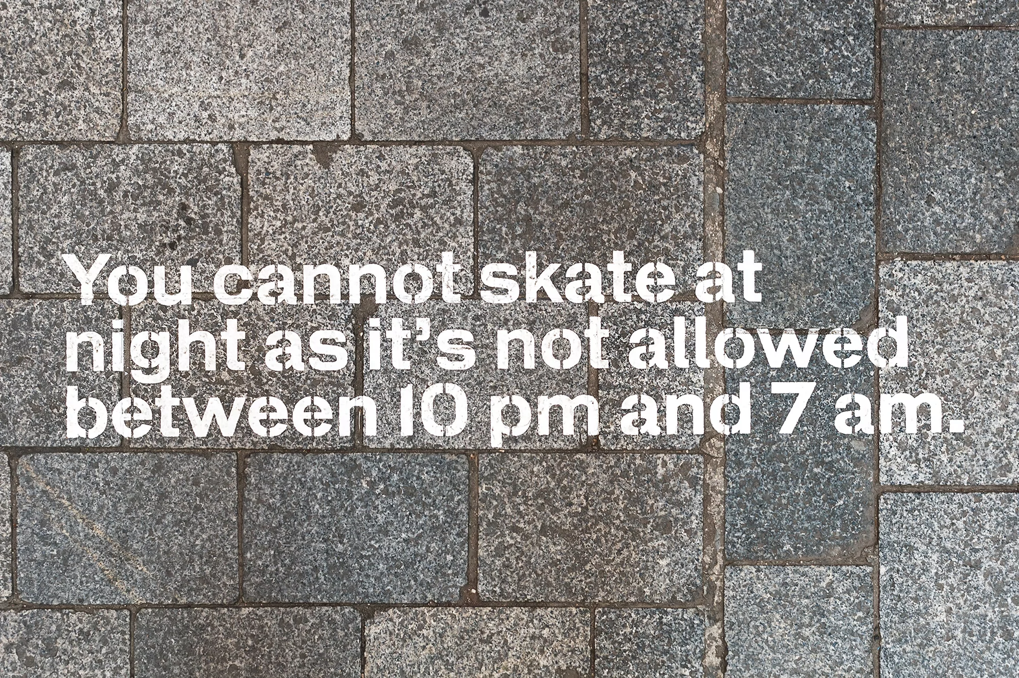 Close-up of gray stone pavement with white stenciled text stating 'You cannot skate at night as it's not allowed between 10 pm and 7 am.'